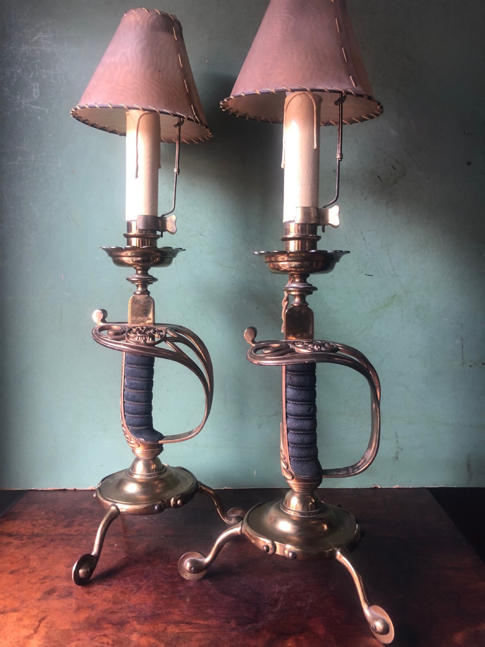 A pair of late nineteenth-century gilded brass candlelamps modelled as cavalry dress swords with basket guards, standing on tripod feet with conical shades. View II
