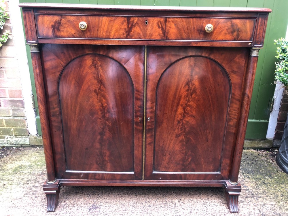 An early nineteenth-century flame mahogany side cabinet with arch-panelled double doors, top drawer, and brass fittings.