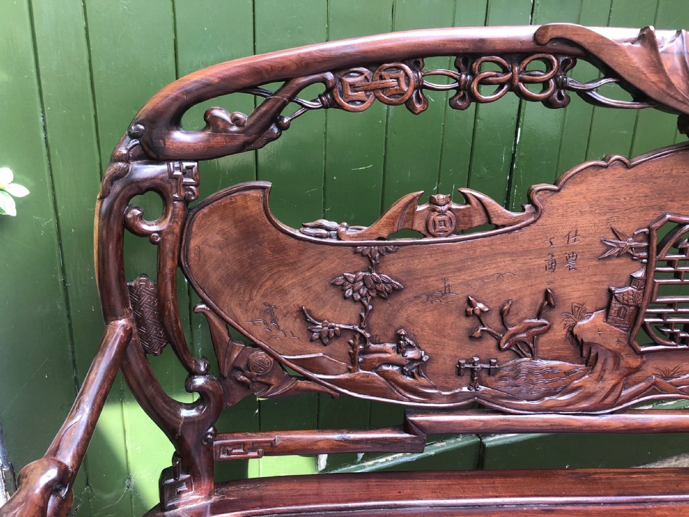 An antique Chinese dark hardwood hall bench featuring detailed relief carvings of bats, landscapes, and traditional character motifs on the backrest. View V