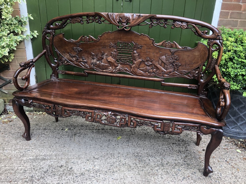 An antique Chinese dark hardwood hall bench featuring detailed relief carvings of bats, landscapes, and traditional character motifs on the backrest. View III