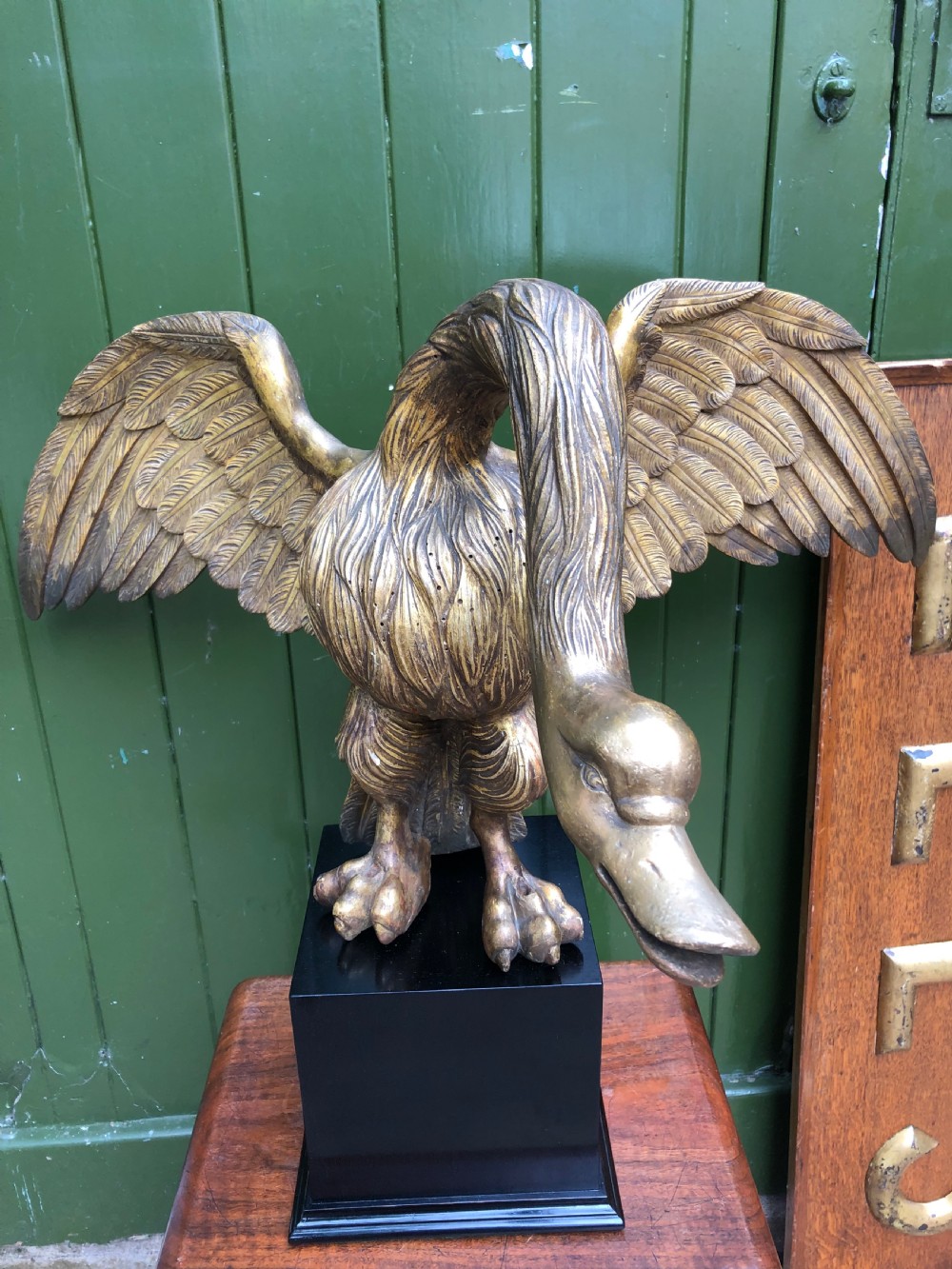 A carved giltwood swan sculpture with outstretched wings and a lowered head mounted on a black square pedestal. View II