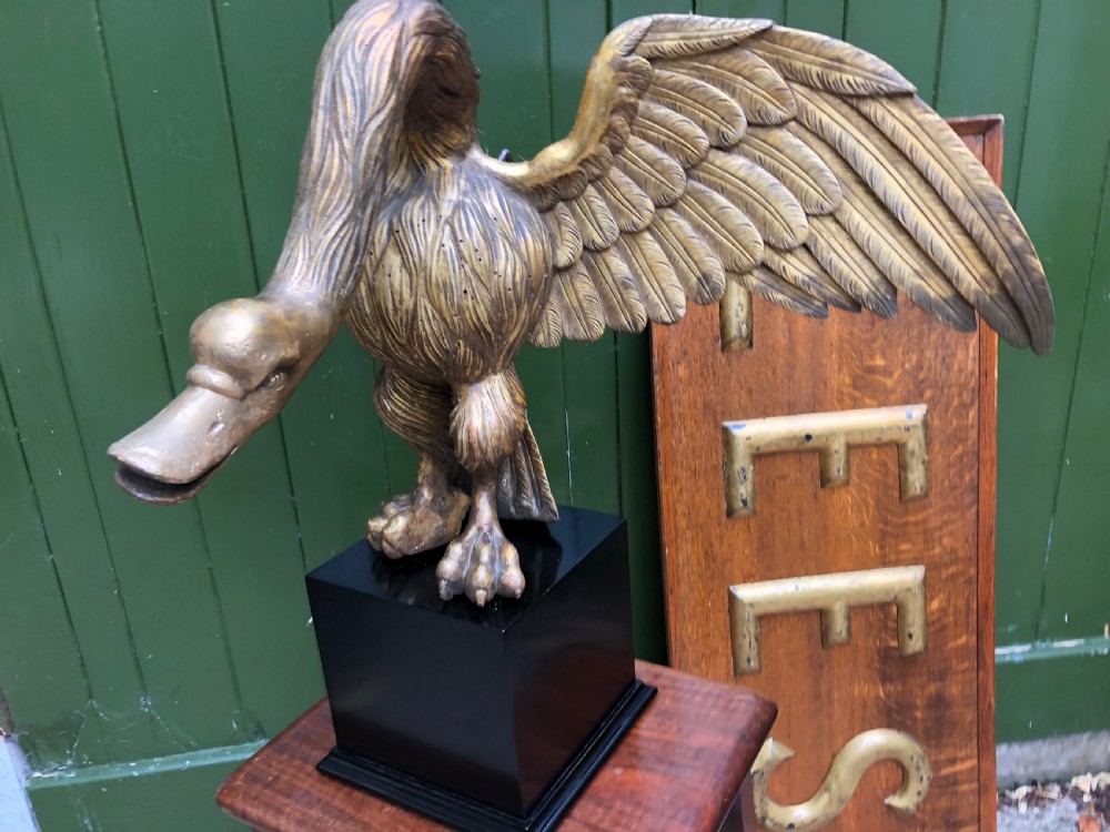 A carved giltwood swan sculpture with outstretched wings and a lowered head mounted on a black square pedestal. View VII