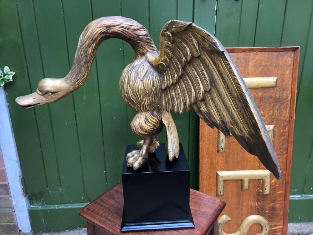 A carved giltwood swan sculpture with outstretched wings and a lowered head mounted on a black square pedestal. View IX