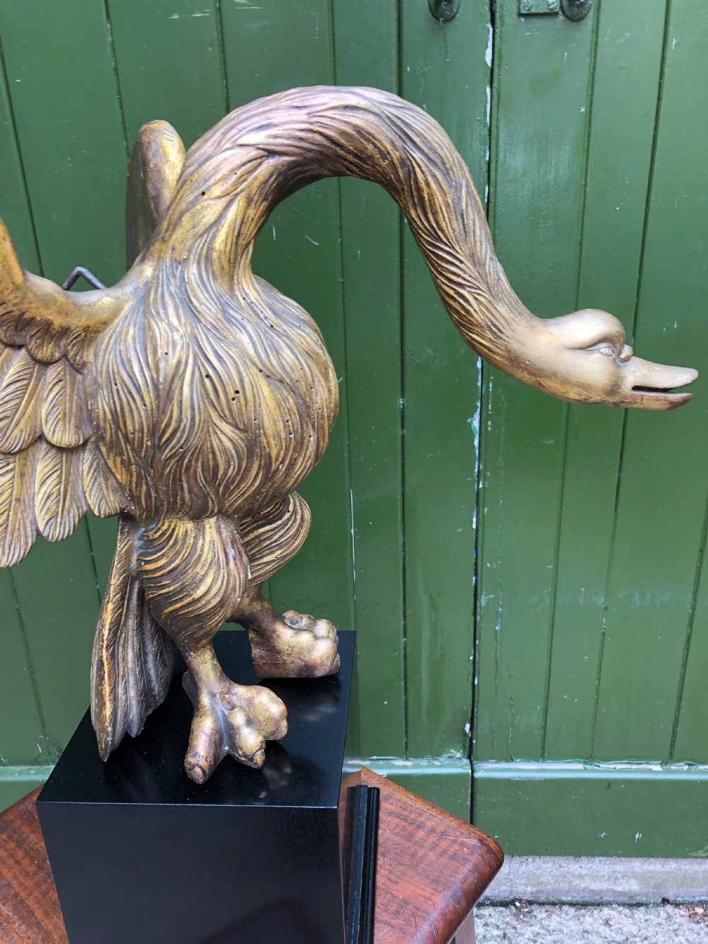 A carved giltwood swan sculpture with outstretched wings and a lowered head mounted on a black square pedestal. View X