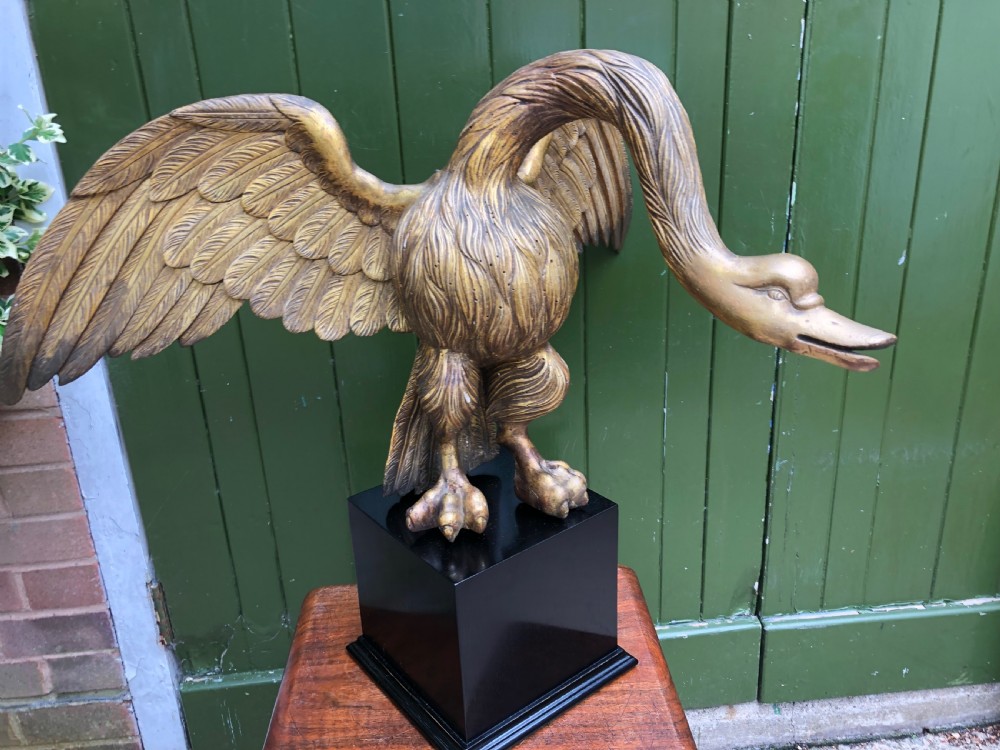 A carved giltwood swan sculpture with outstretched wings and a lowered head mounted on a black square pedestal. View XVI