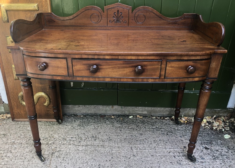 An early nineteenth-century Regency mahogany side table featuring a carved gallery back, three drawers with turned knobs, and turned legs resting on brass castors.