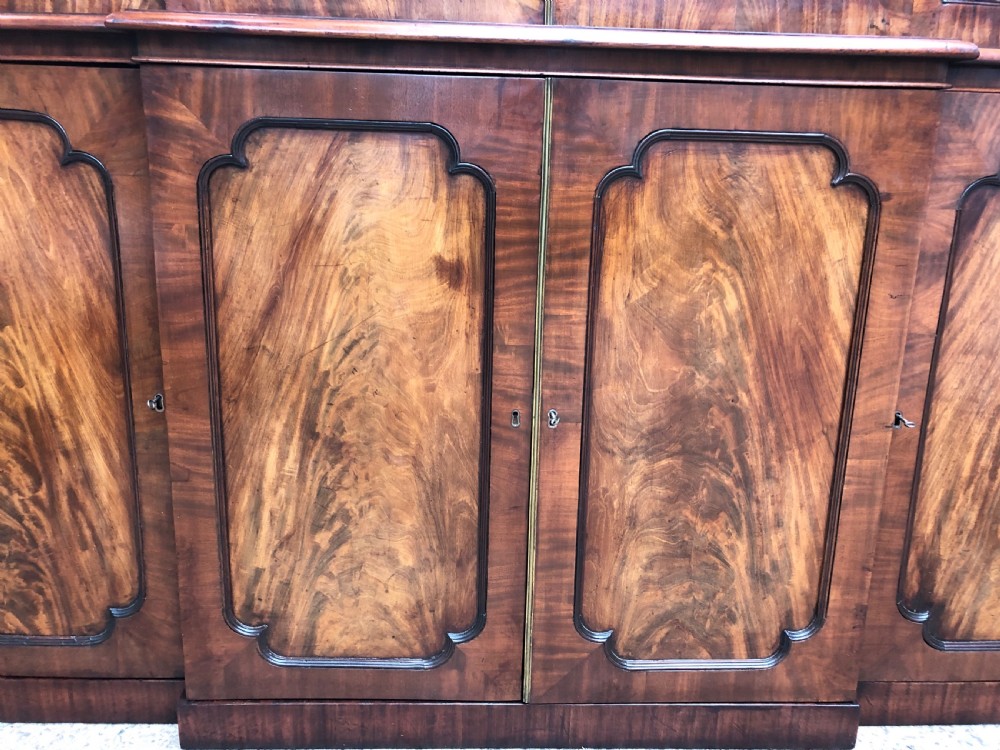 An early 19th-century George IV flame mahogany breakfront library bookcase featuring four upper glazed doors with astragal glazing bars above four fielded panel lower cupboards. View X