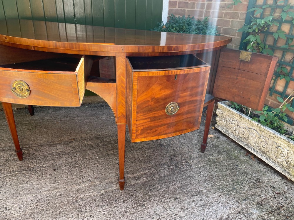 A late 18th-century George III mahogany demi-lune sideboard featuring inlaid stringing, a central drawer, curved side doors, and tapered square legs. View X