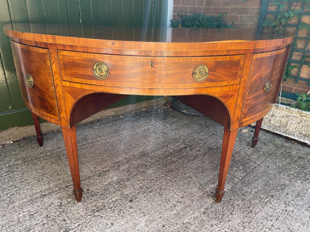 A late 18th-century George III mahogany demi-lune sideboard featuring inlaid stringing, a central drawer, curved side doors, and tapered square legs. View XX