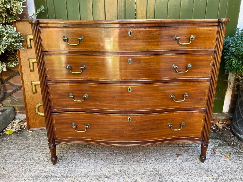 A George III Sheraton period mahogany chest of drawers featuring a curved serpentine front, four graduated drawers, brass swan-neck handles, and reeded corner columns. View II
