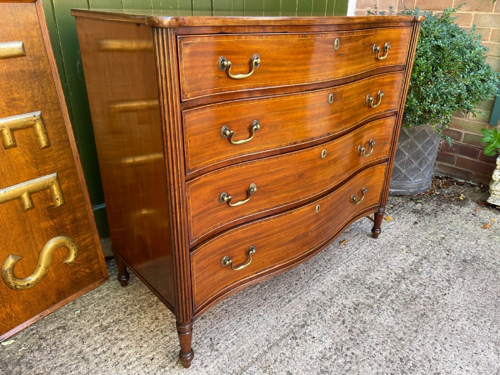 A George III Sheraton period mahogany chest of drawers featuring a curved serpentine front, four graduated drawers, brass swan-neck handles, and reeded corner columns. View III