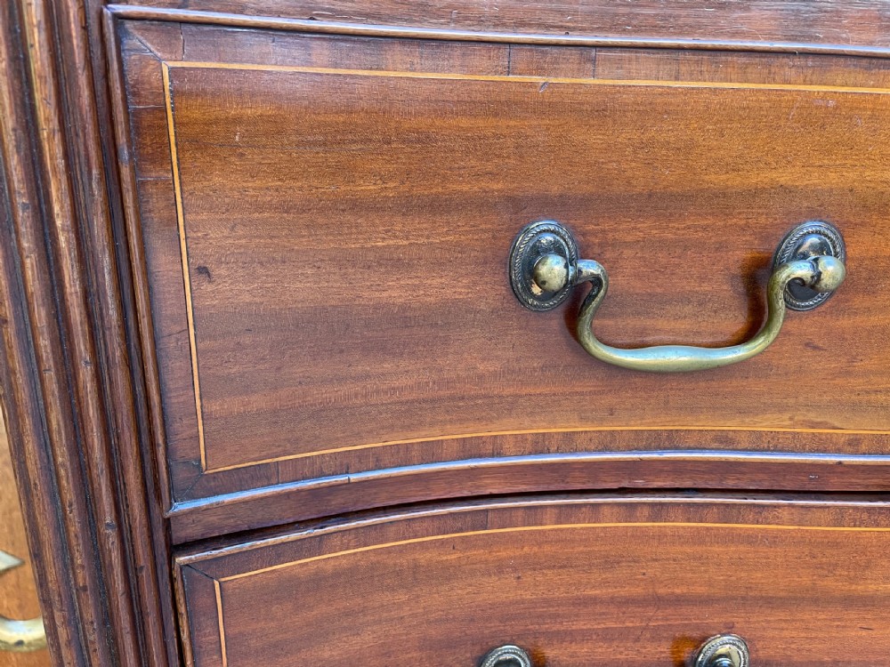 A George III Sheraton period mahogany chest of drawers featuring a curved serpentine front, four graduated drawers, brass swan-neck handles, and reeded corner columns. View VI