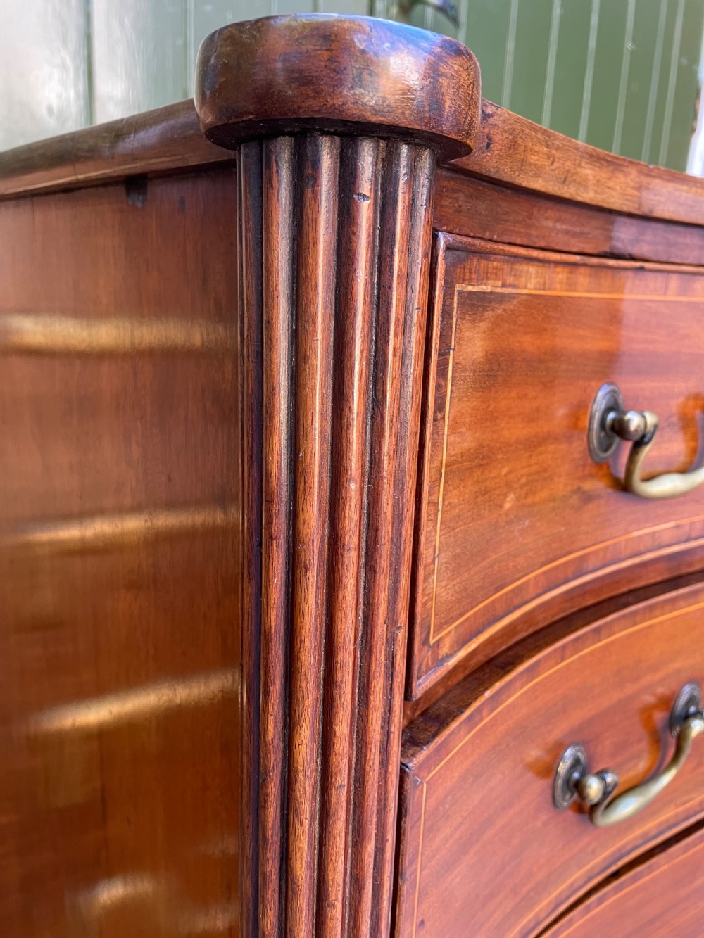 A George III Sheraton period mahogany chest of drawers featuring a curved serpentine front, four graduated drawers, brass swan-neck handles, and reeded corner columns. View VII
