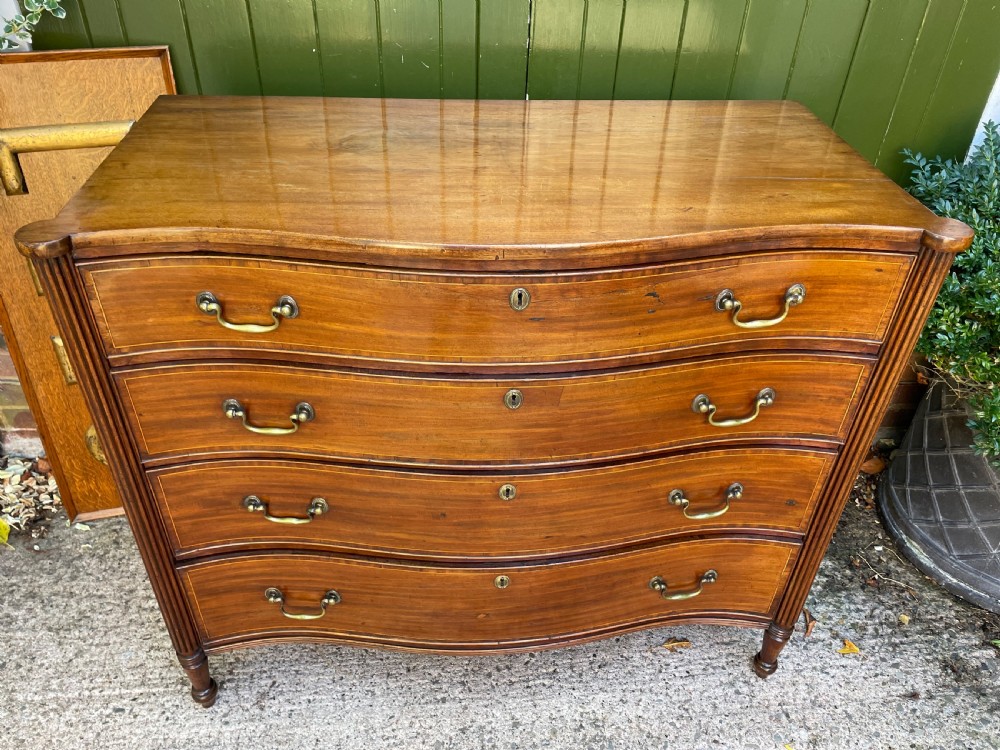 A George III Sheraton period mahogany chest of drawers featuring a curved serpentine front, four graduated drawers, brass swan-neck handles, and reeded corner columns. View VIII