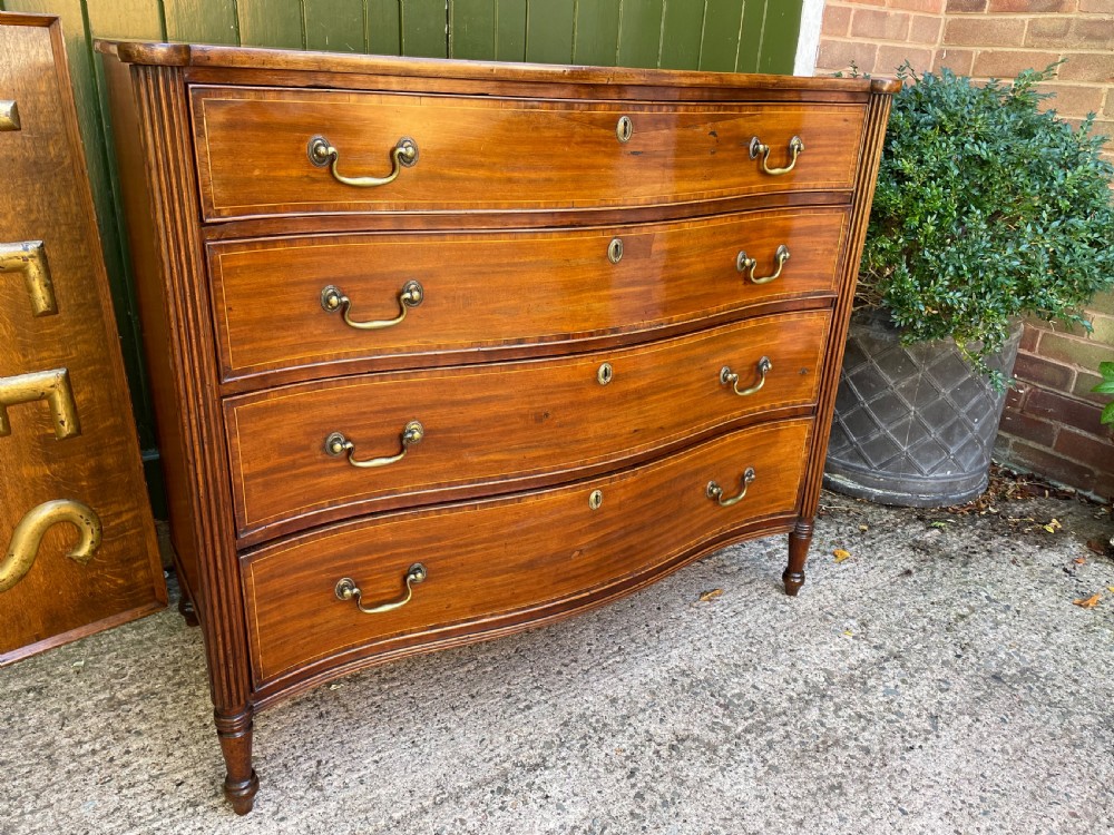 A George III Sheraton period mahogany chest of drawers featuring a curved serpentine front, four graduated drawers, brass swan-neck handles, and reeded corner columns. View X