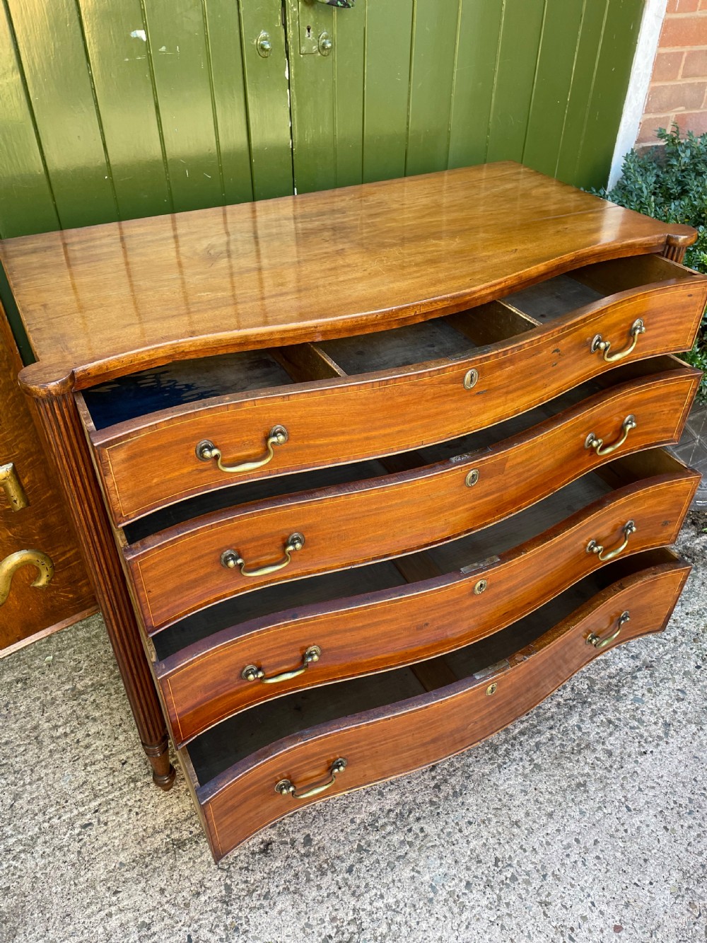 A George III Sheraton period mahogany chest of drawers featuring a curved serpentine front, four graduated drawers, brass swan-neck handles, and reeded corner columns. View XI