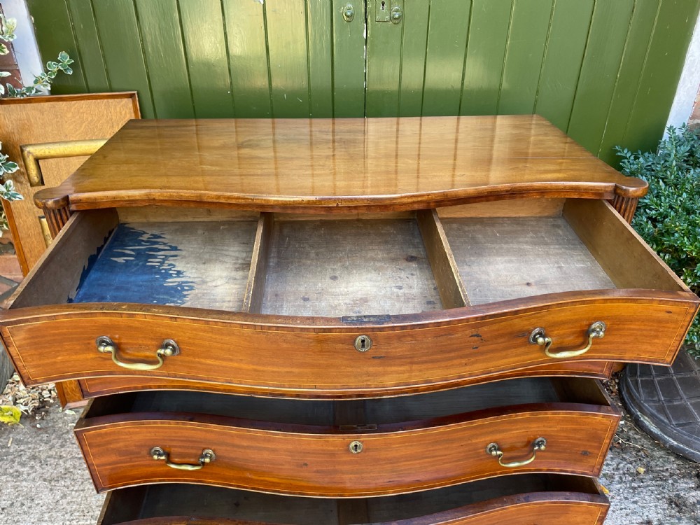 A George III Sheraton period mahogany chest of drawers featuring a curved serpentine front, four graduated drawers, brass swan-neck handles, and reeded corner columns. View XII