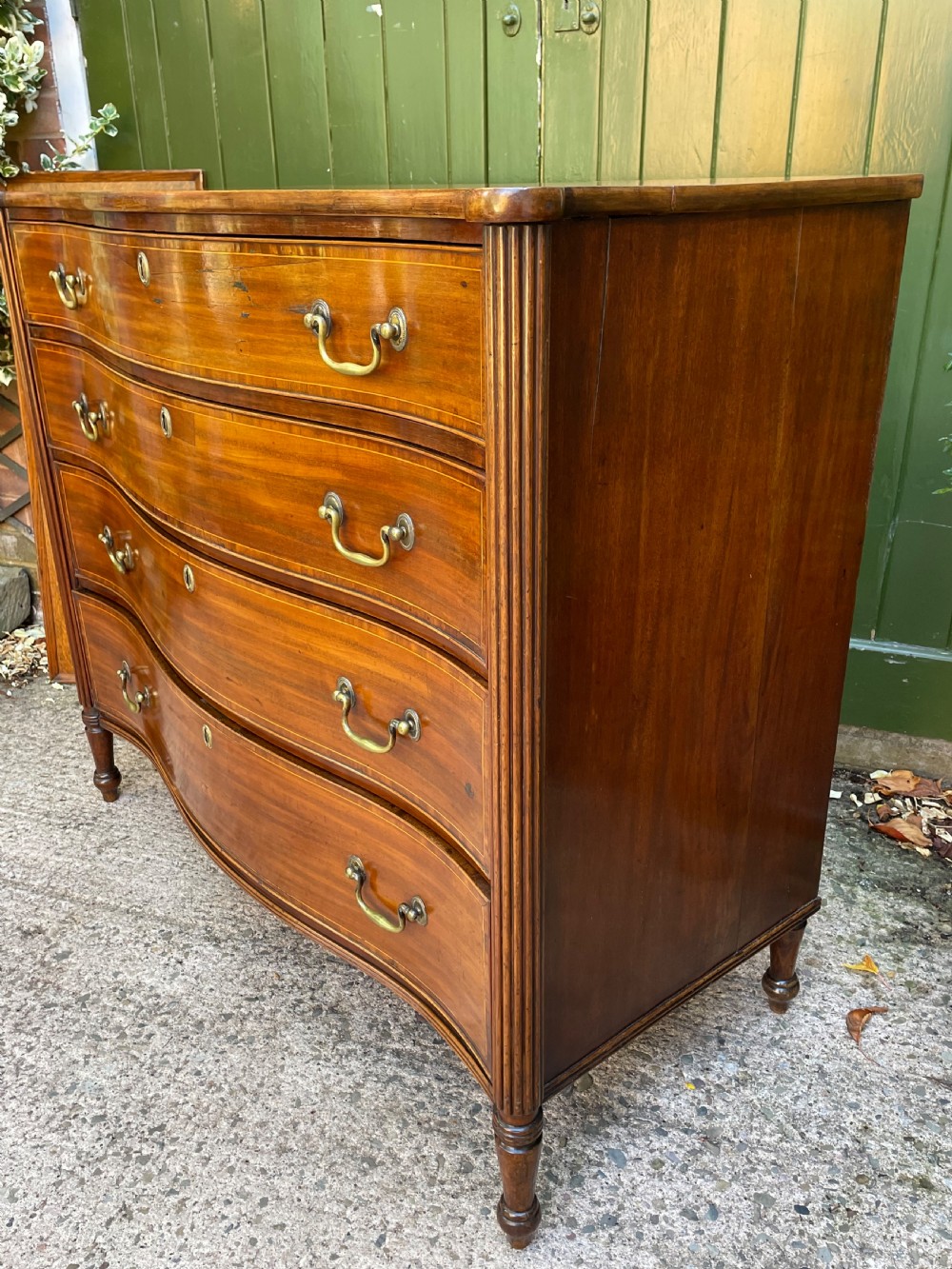 A George III Sheraton period mahogany chest of drawers featuring a curved serpentine front, four graduated drawers, brass swan-neck handles, and reeded corner columns. View XV