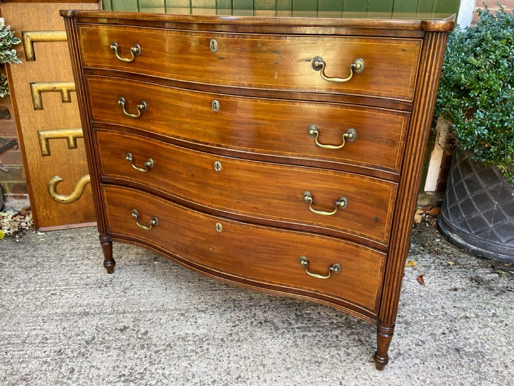 A George III Sheraton period mahogany chest of drawers featuring a curved serpentine front, four graduated drawers, brass swan-neck handles, and reeded corner columns. View XVI