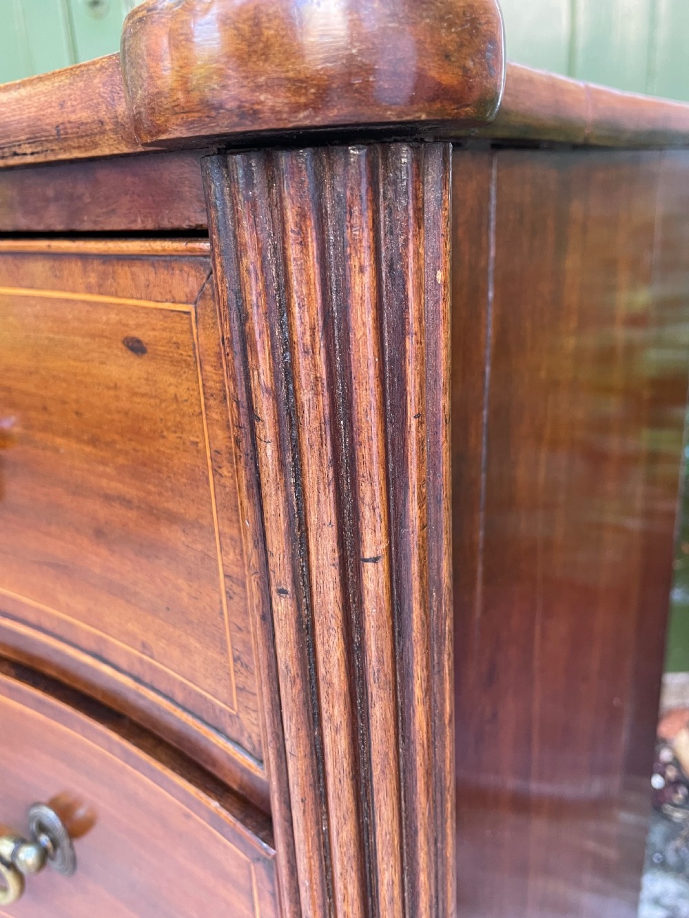 A George III Sheraton period mahogany chest of drawers featuring a curved serpentine front, four graduated drawers, brass swan-neck handles, and reeded corner columns. View XVII
