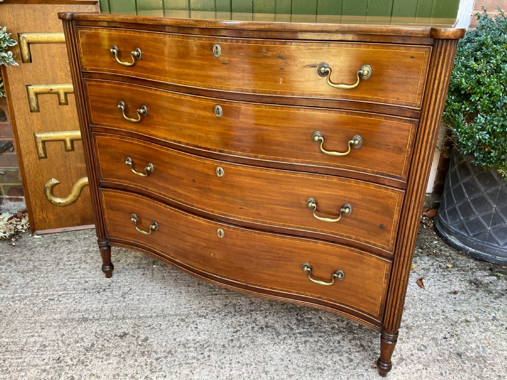 A George III Sheraton period mahogany chest of drawers featuring a curved serpentine front, four graduated drawers, brass swan-neck handles, and reeded corner columns. View XIX