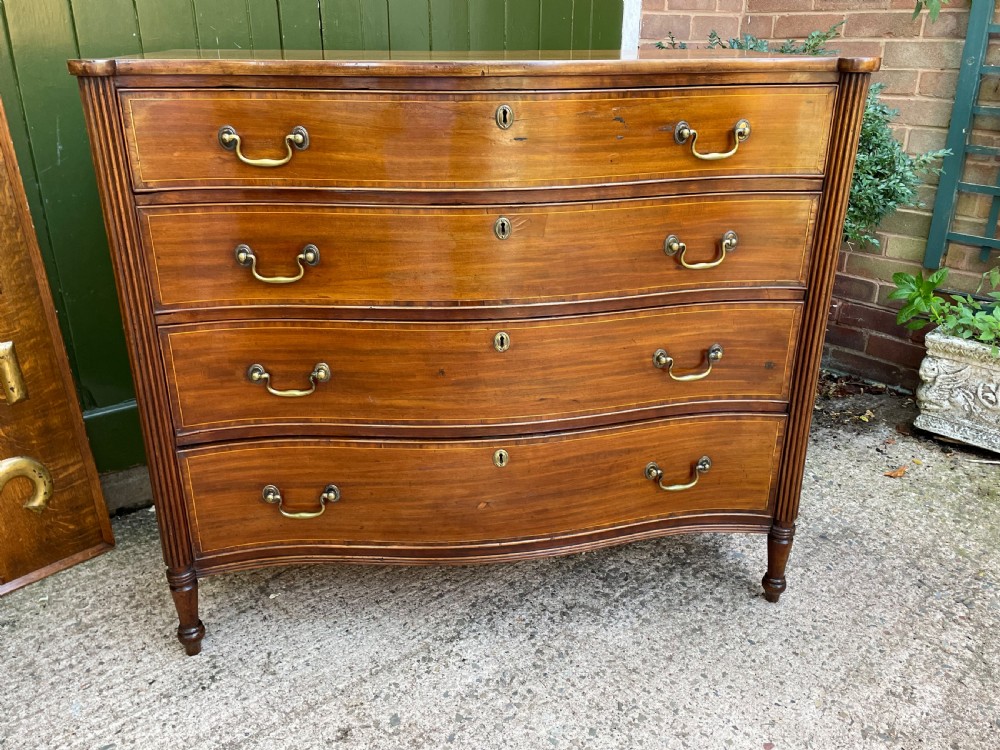 A George III Sheraton period mahogany chest of drawers featuring a curved serpentine front, four graduated drawers, brass swan-neck handles, and reeded corner columns. View XXI