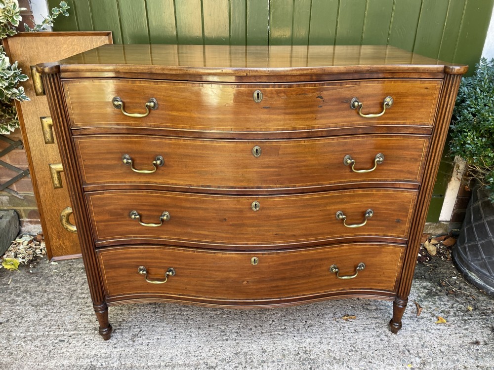 A George III Sheraton period mahogany chest of drawers featuring a curved serpentine front, four graduated drawers, brass swan-neck handles, and reeded corner columns.