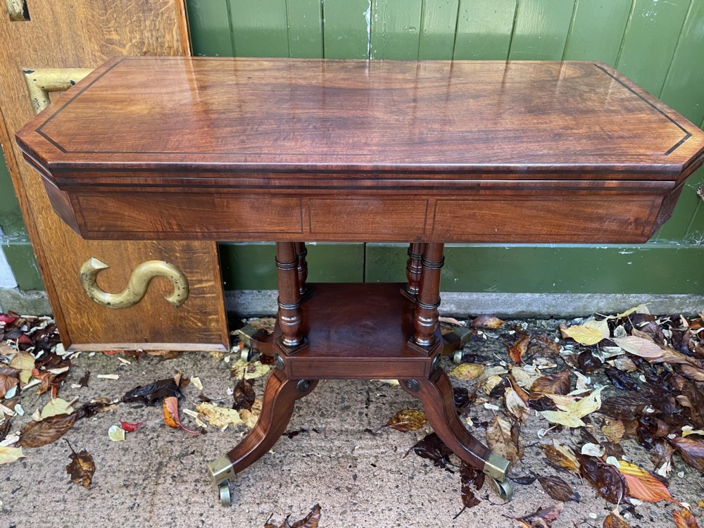An early 19th-century Regency mahogany fold-over card table featuring turned column supports, a square platform base, and four splayed sabre legs with brass castors. View II
