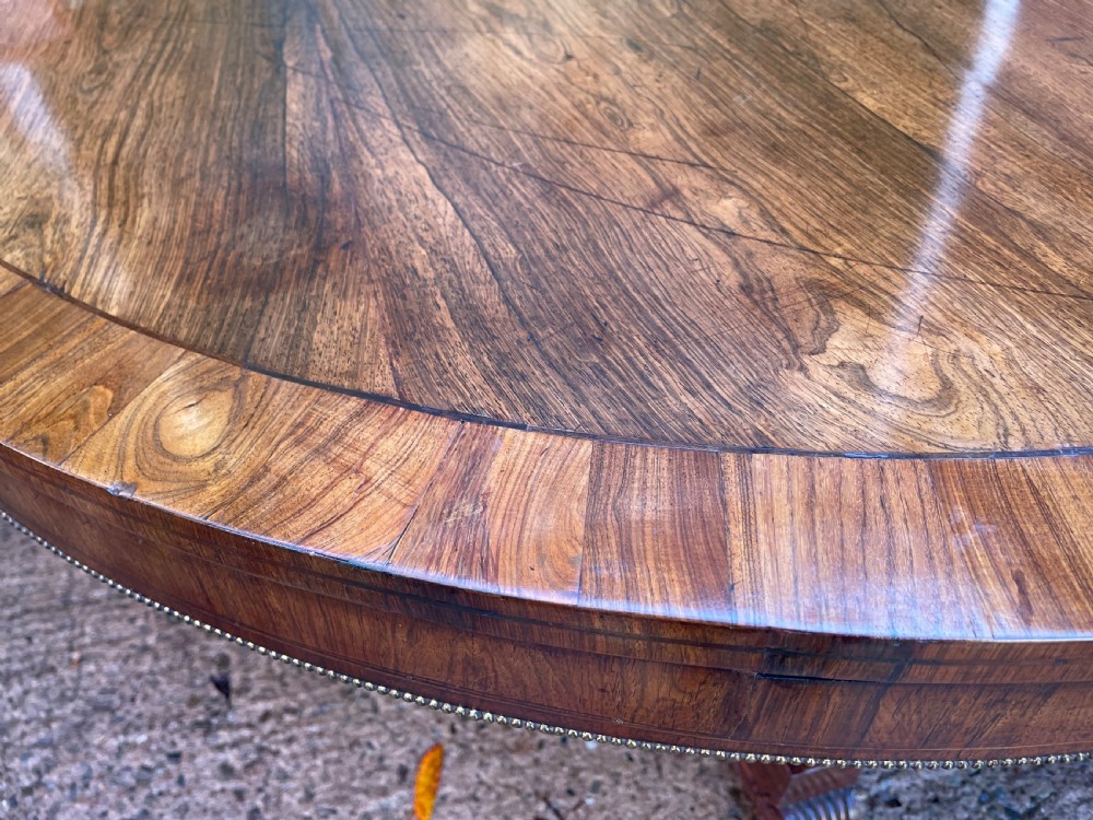 An early nineteenth-century circular Regency rosewood centre table featuring a crossbanded border, displayed in a tilted position on a pedestal base with castors. View XIX