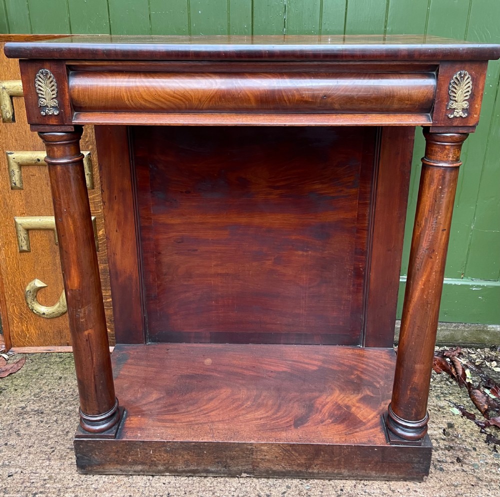 Early 19th-century George IV mahogany console table featuring cylindrical front columns, brass mounts, a plinth base, and a flame mahogany backdrop.