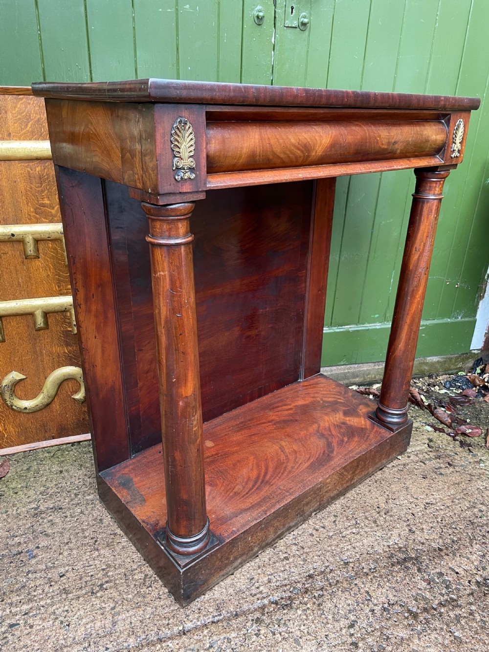 Early 19th-century George IV mahogany console table featuring cylindrical front columns, brass mounts, a plinth base, and a flame mahogany backdrop. View XII