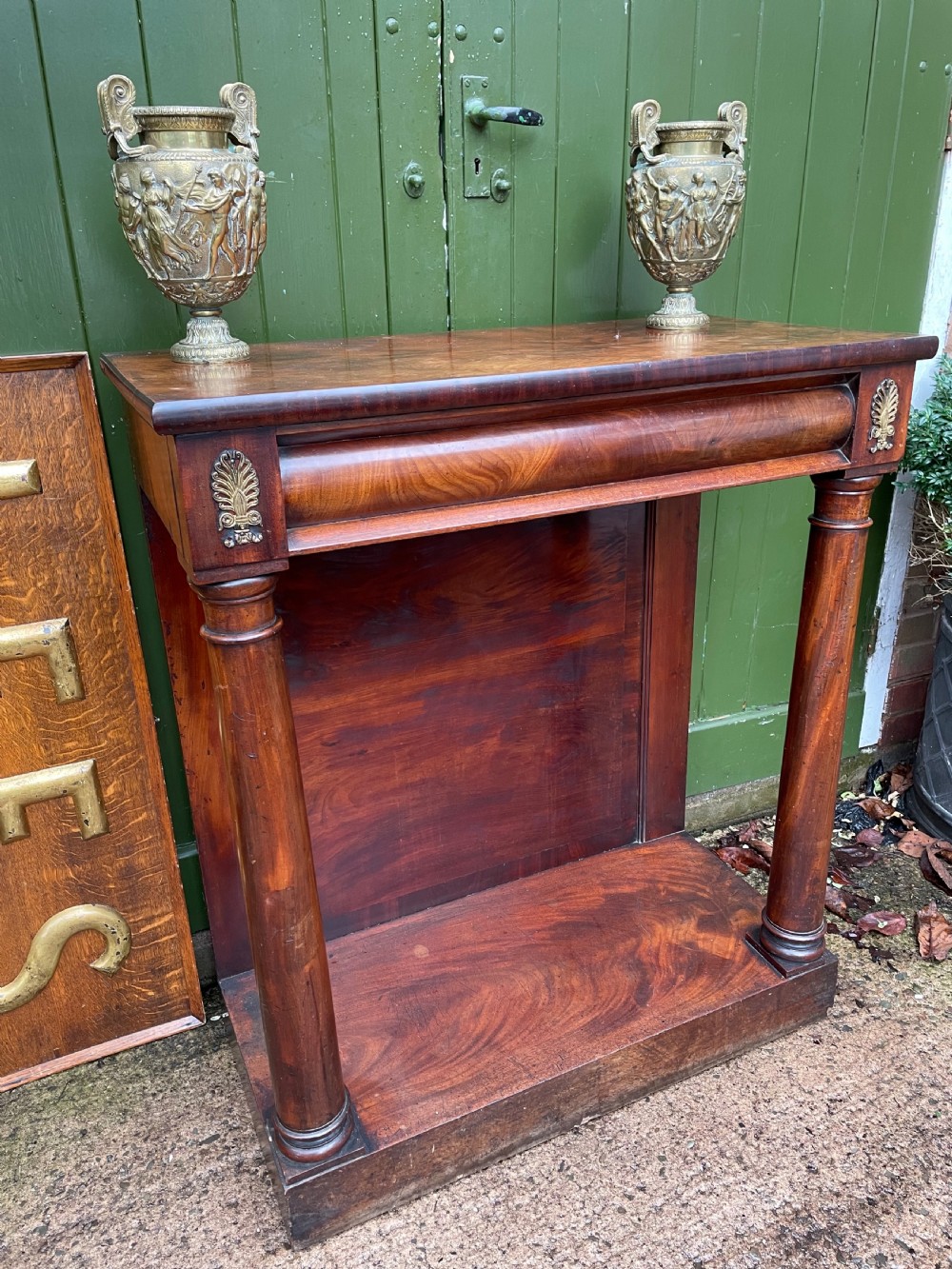 Early 19th-century George IV mahogany console table featuring cylindrical front columns, brass mounts, a plinth base, and a flame mahogany backdrop. View XV