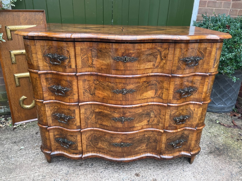 A mid-18th-century German Baroque walnut commode featuring a shaped serpentine front, four long drawers with figured wood veneers, and ornate cast brass handles. View XVI