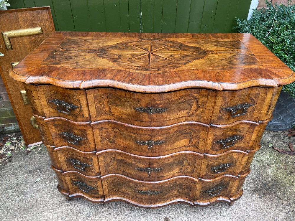 A mid-18th-century German Baroque walnut commode featuring a shaped serpentine front, four long drawers with figured wood veneers, and ornate cast brass handles. View XVIII
