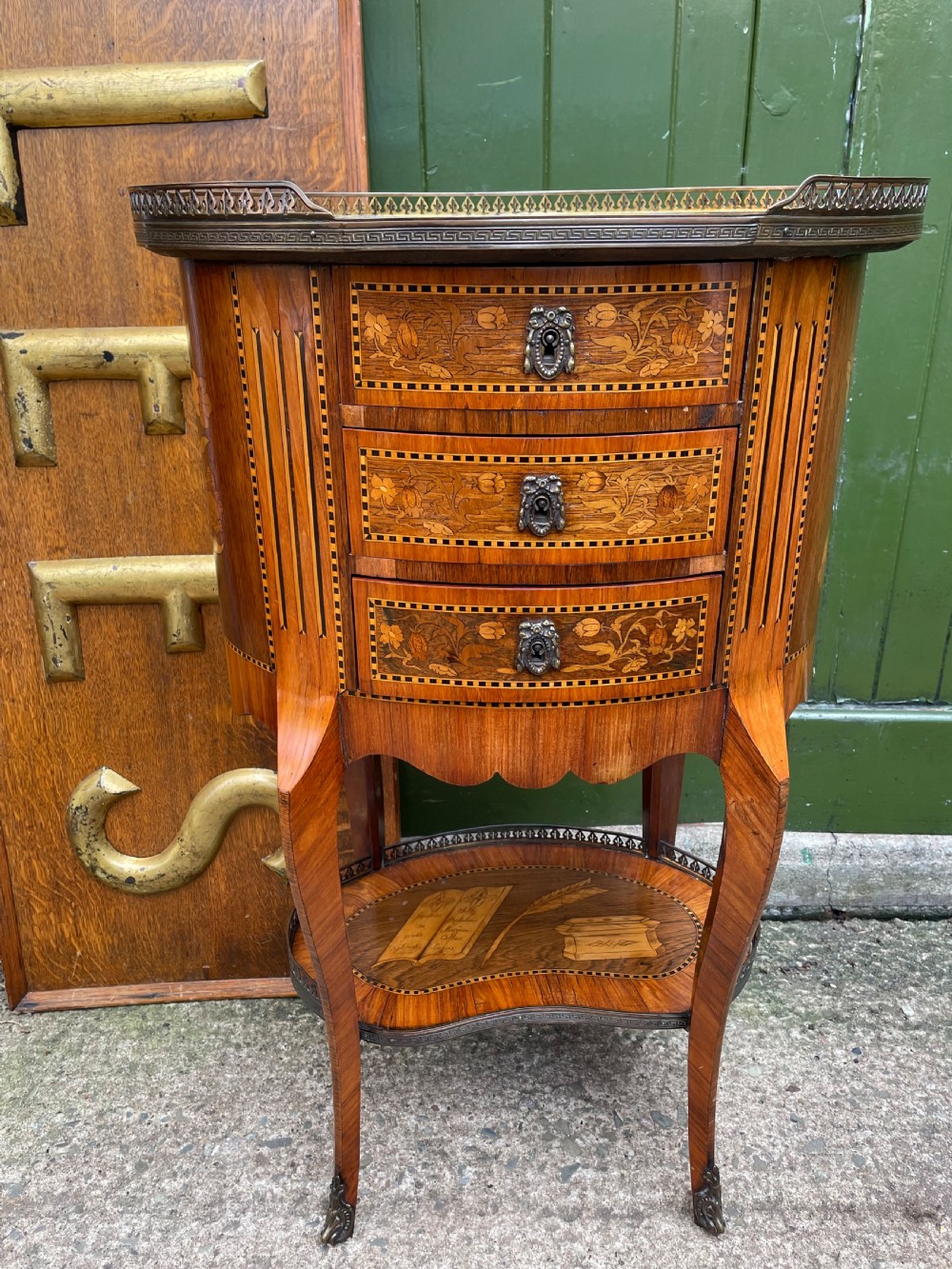 A late nineteenth-century French oval marquetry table featuring three drawers with floral inlay, a pierced brass gallery top, and a lower shelf decorated with a marquetry book design.
