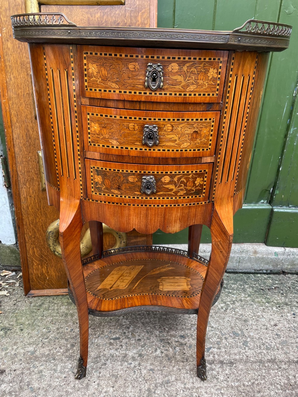 A late nineteenth-century French oval marquetry table featuring three drawers with floral inlay, a pierced brass gallery top, and a lower shelf decorated with a marquetry book design. View X