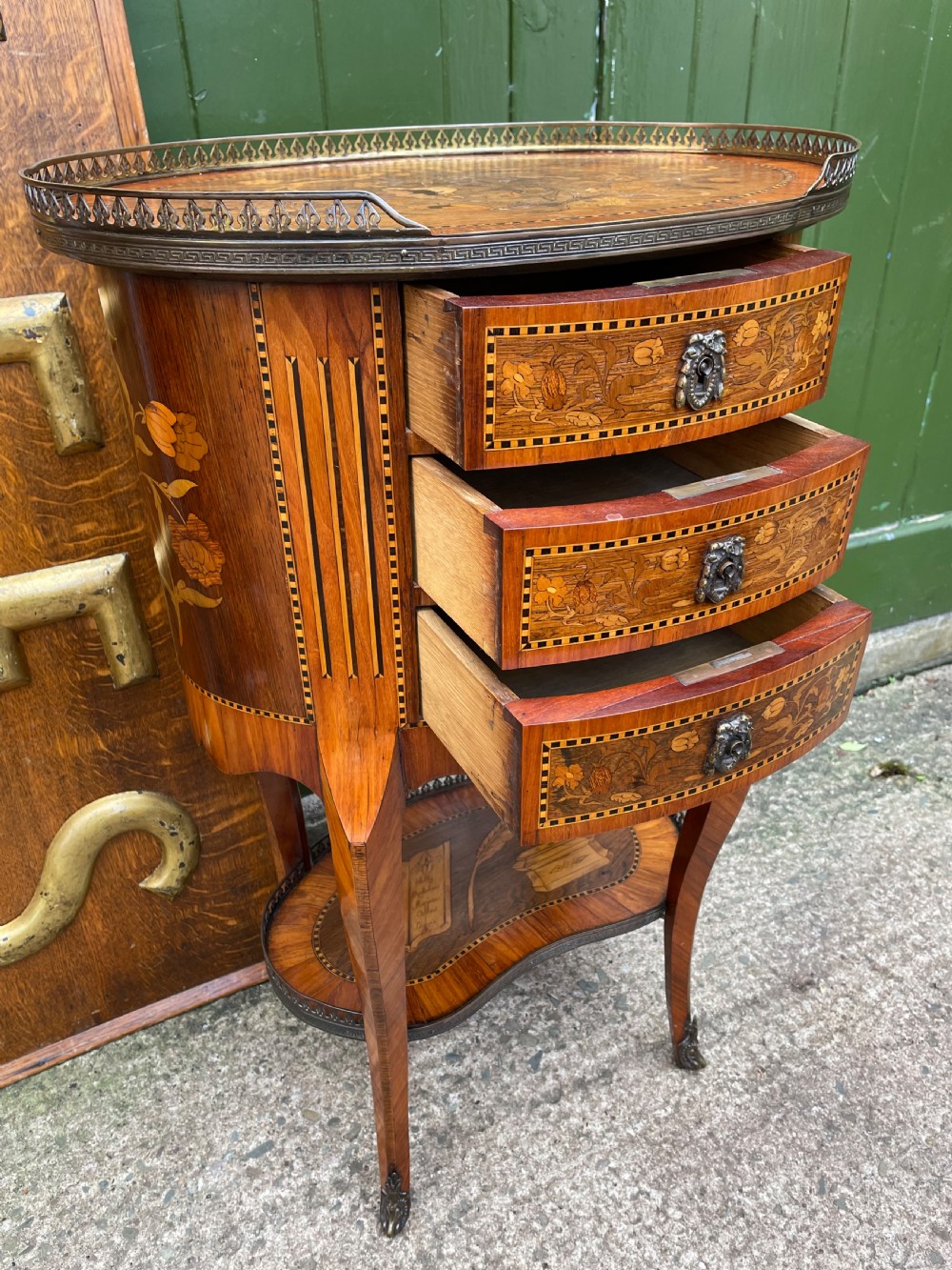 A late nineteenth-century French oval marquetry table featuring three drawers with floral inlay, a pierced brass gallery top, and a lower shelf decorated with a marquetry book design. View XIII