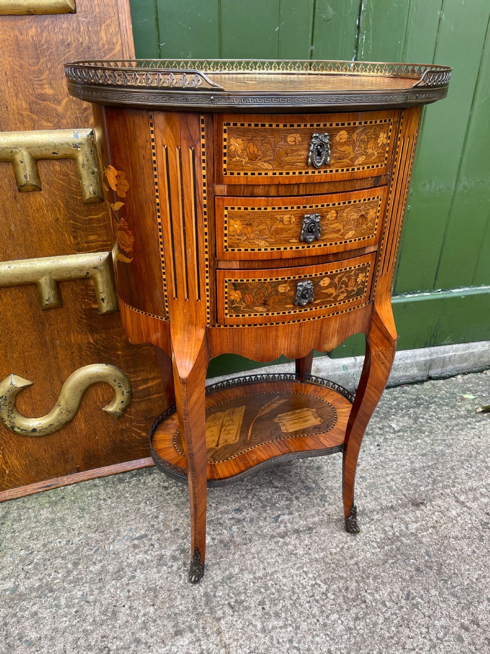 A late nineteenth-century French oval marquetry table featuring three drawers with floral inlay, a pierced brass gallery top, and a lower shelf decorated with a marquetry book design. View XV