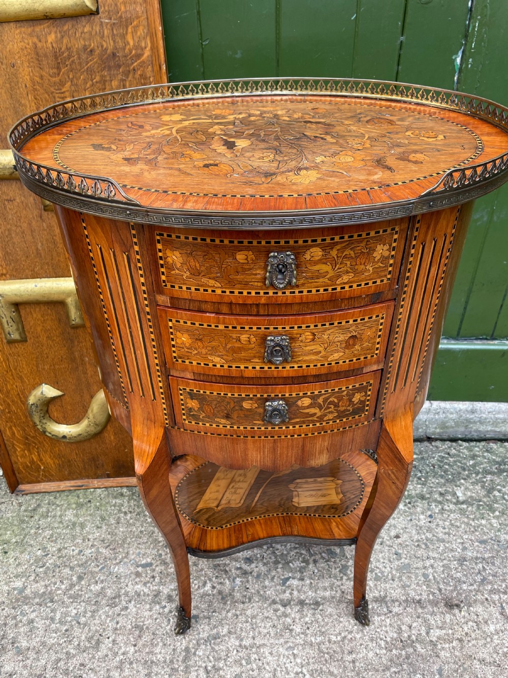 A late nineteenth-century French oval marquetry table featuring three drawers with floral inlay, a pierced brass gallery top, and a lower shelf decorated with a marquetry book design. View XIX