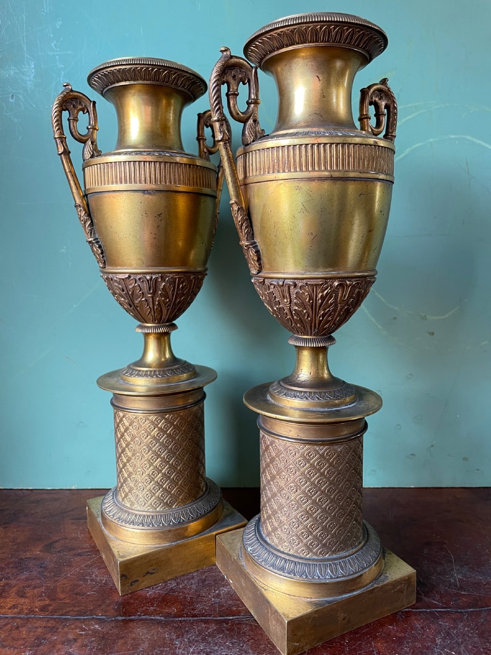 A pair of early 19th-century French gilt-bronze Neoclassical urns featuring scrolled handles, acanthus leaf relief, and diaper-patterned cylindrical plinths. View V
