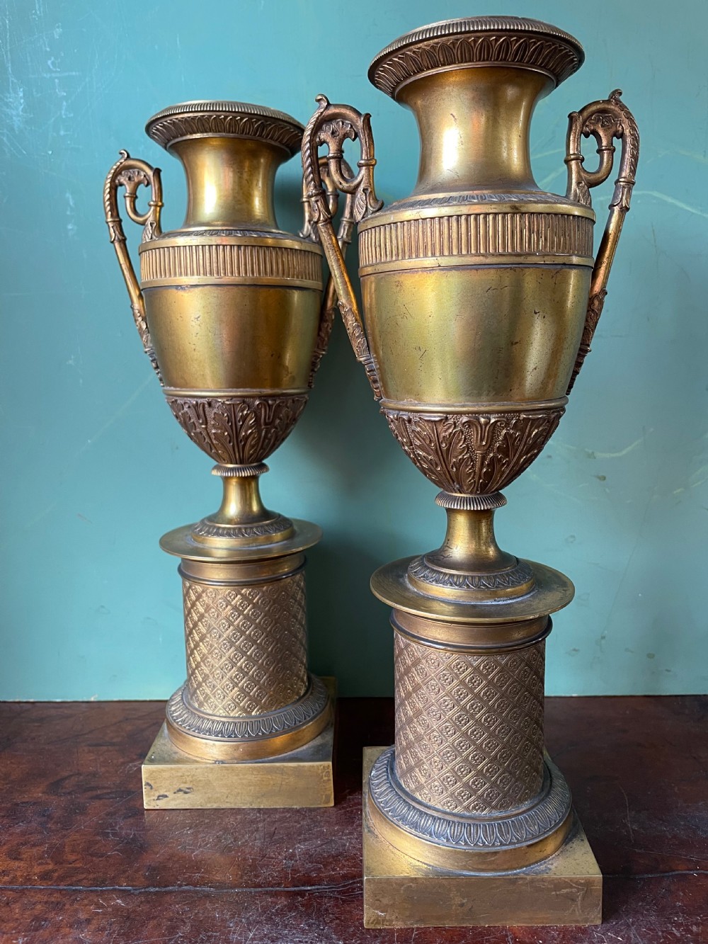 A pair of early 19th-century French gilt-bronze Neoclassical urns featuring scrolled handles, acanthus leaf relief, and diaper-patterned cylindrical plinths. View VI