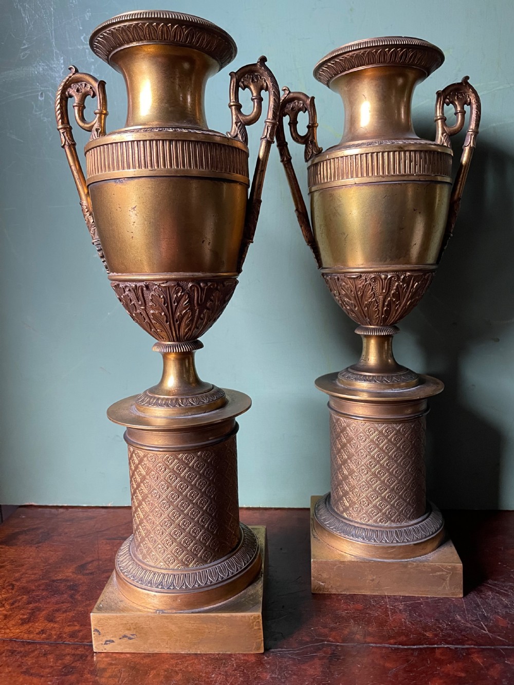 A pair of early 19th-century French gilt-bronze Neoclassical urns featuring scrolled handles, acanthus leaf relief, and diaper-patterned cylindrical plinths. View VII