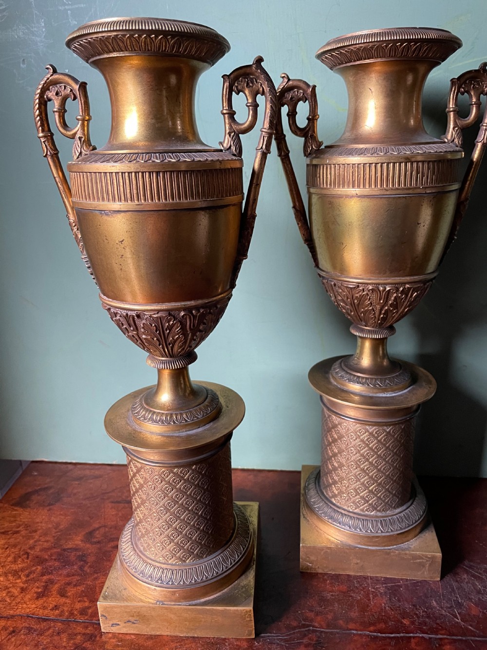 A pair of early 19th-century French gilt-bronze Neoclassical urns featuring scrolled handles, acanthus leaf relief, and diaper-patterned cylindrical plinths. View VIII