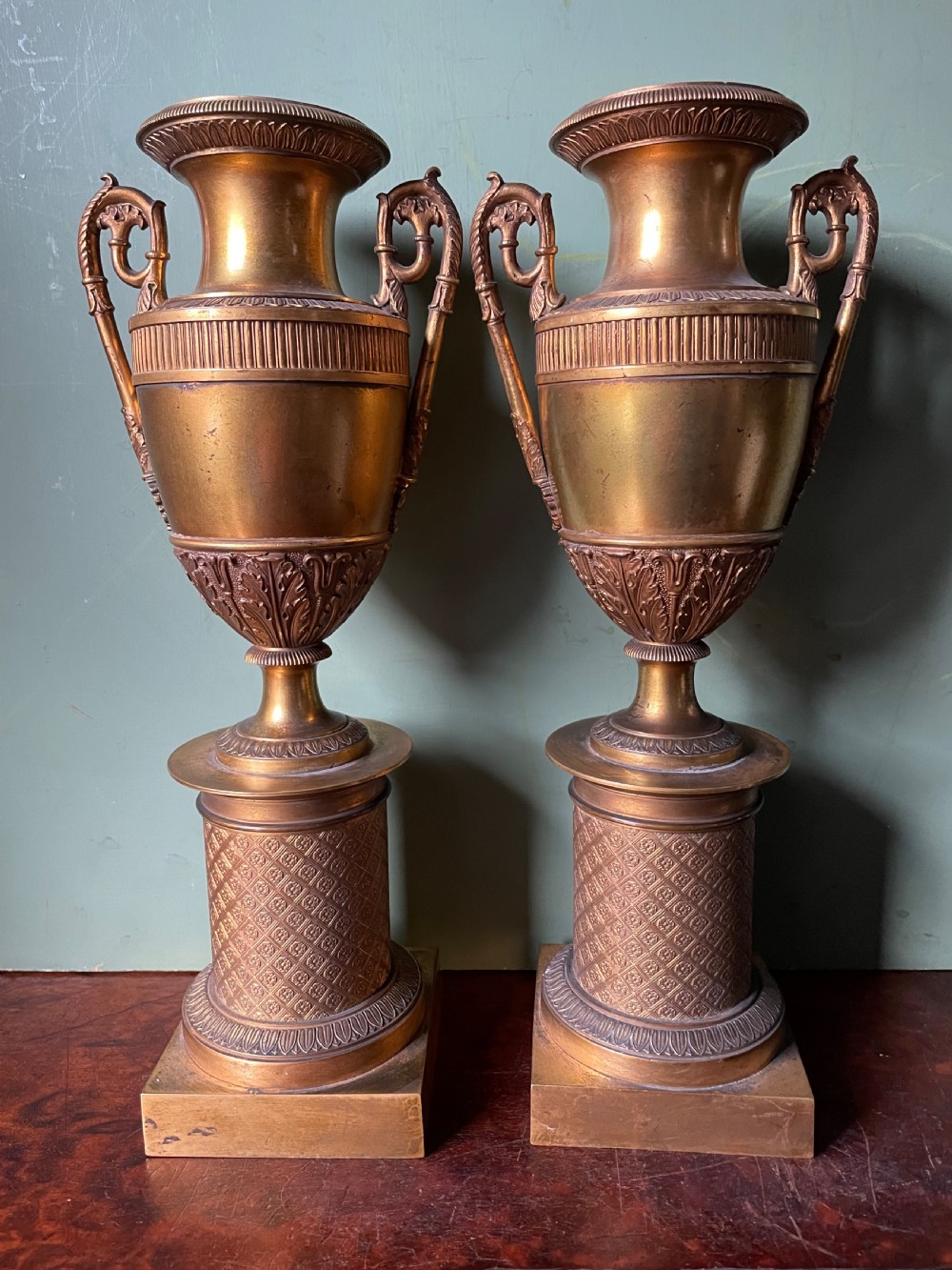 A pair of early 19th-century French gilt-bronze Neoclassical urns featuring scrolled handles, acanthus leaf relief, and diaper-patterned cylindrical plinths. View IX