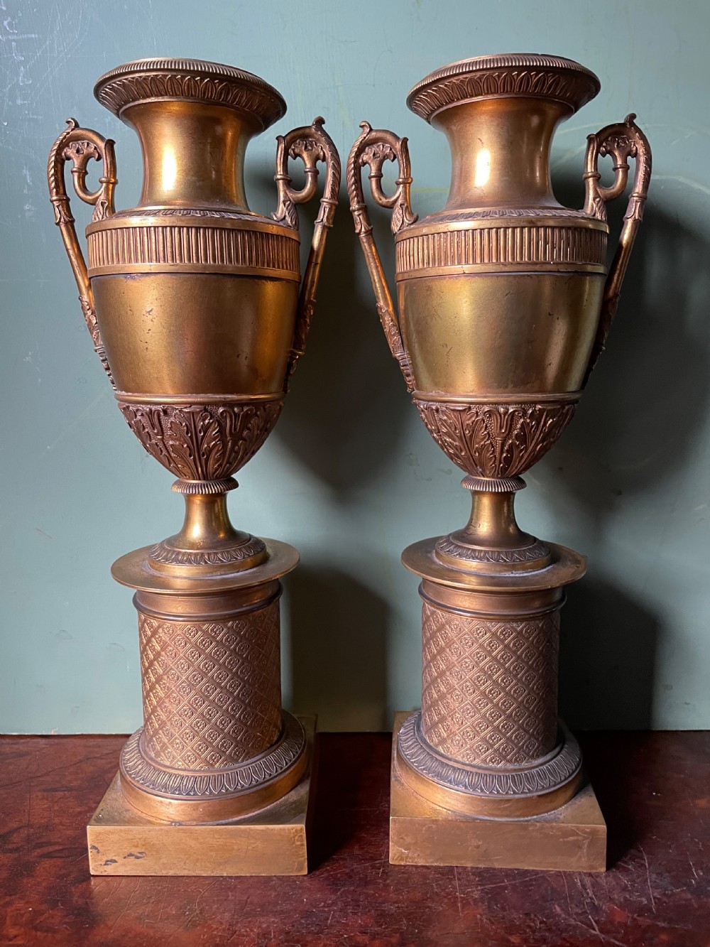 A pair of early 19th-century French gilt-bronze Neoclassical urns featuring scrolled handles, acanthus leaf relief, and diaper-patterned cylindrical plinths. View X