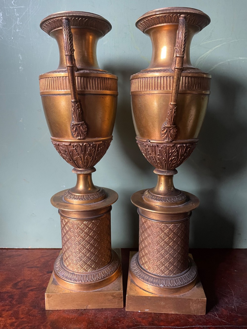 A pair of early 19th-century French gilt-bronze Neoclassical urns featuring scrolled handles, acanthus leaf relief, and diaper-patterned cylindrical plinths. View XVI