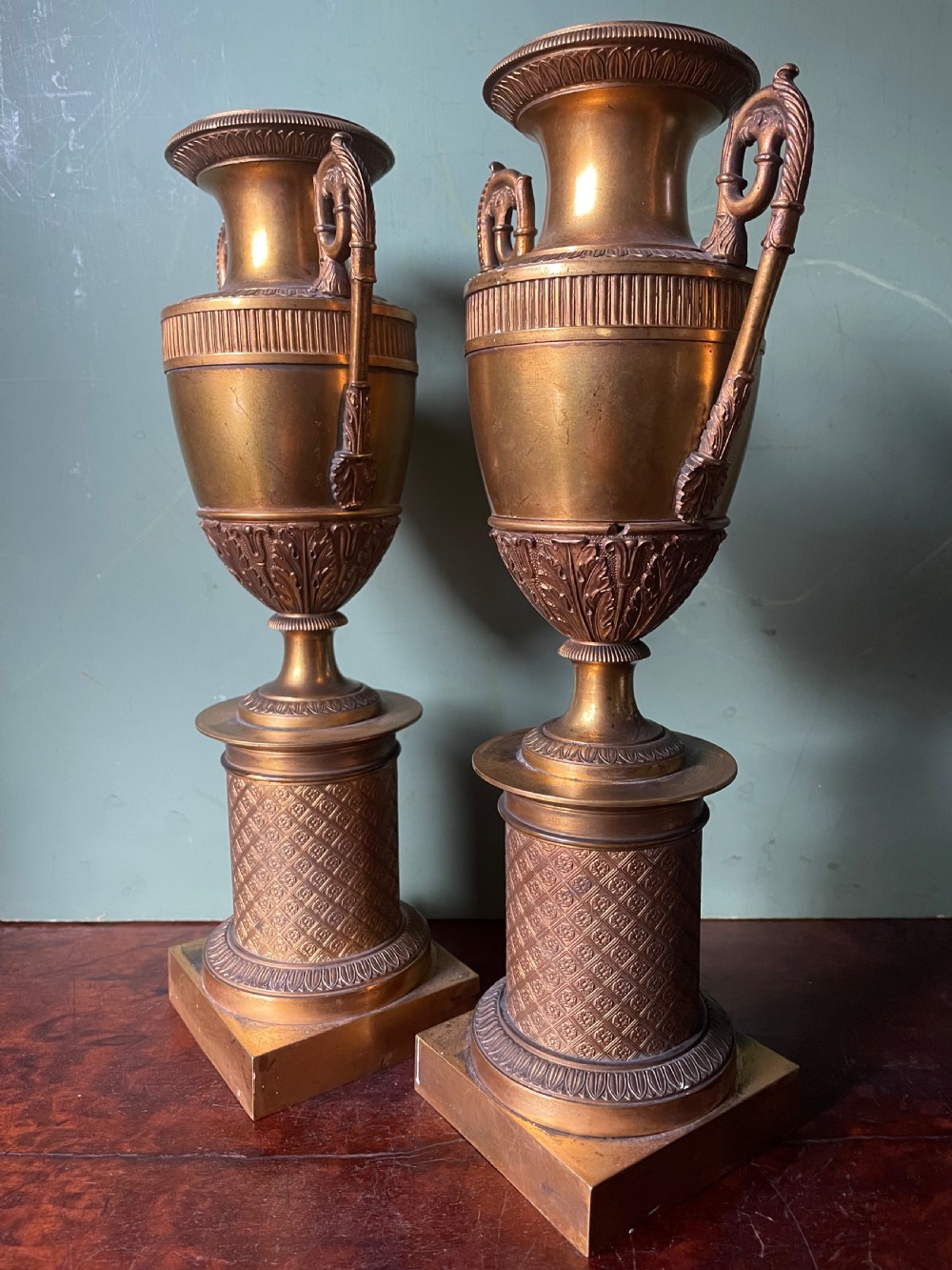 A pair of early 19th-century French gilt-bronze Neoclassical urns featuring scrolled handles, acanthus leaf relief, and diaper-patterned cylindrical plinths. View XVII