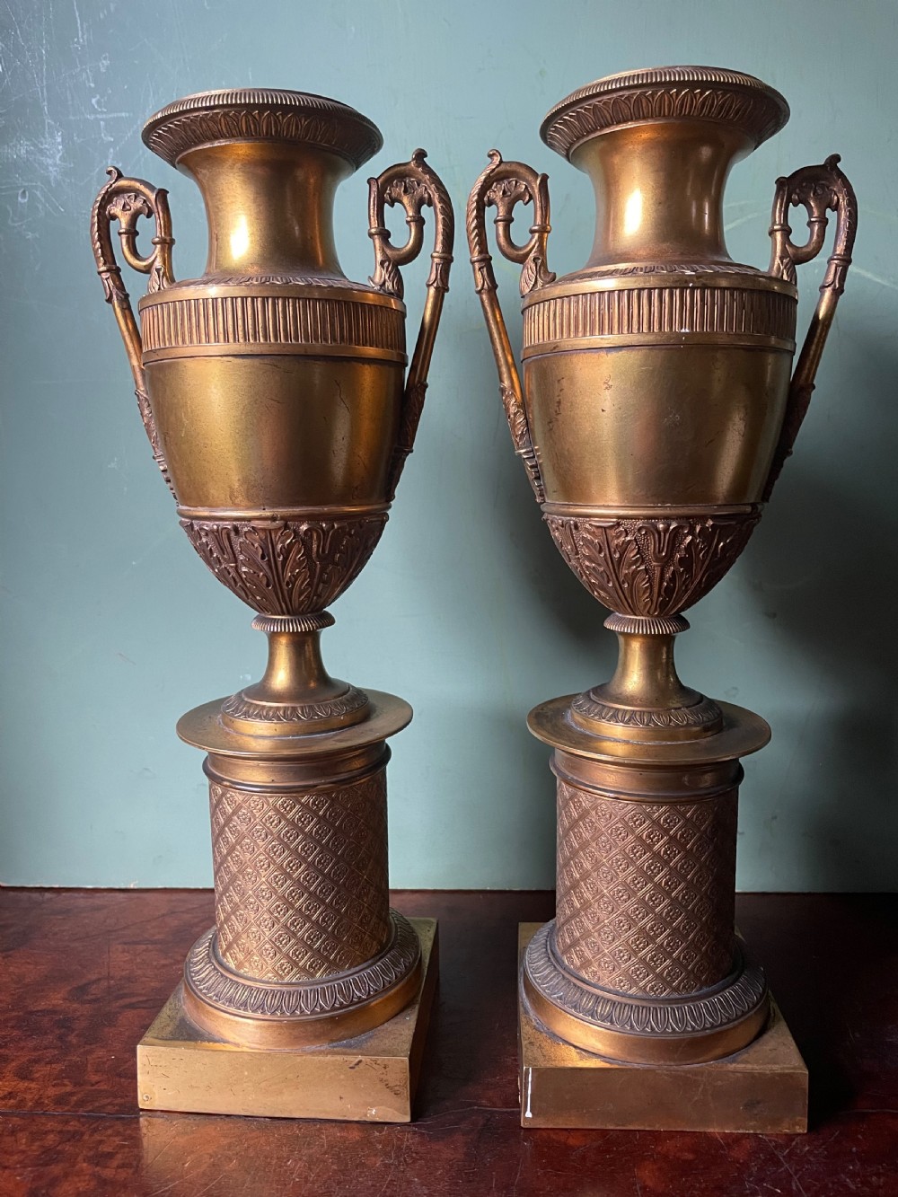A pair of early 19th-century French gilt-bronze Neoclassical urns featuring scrolled handles, acanthus leaf relief, and diaper-patterned cylindrical plinths. View XX