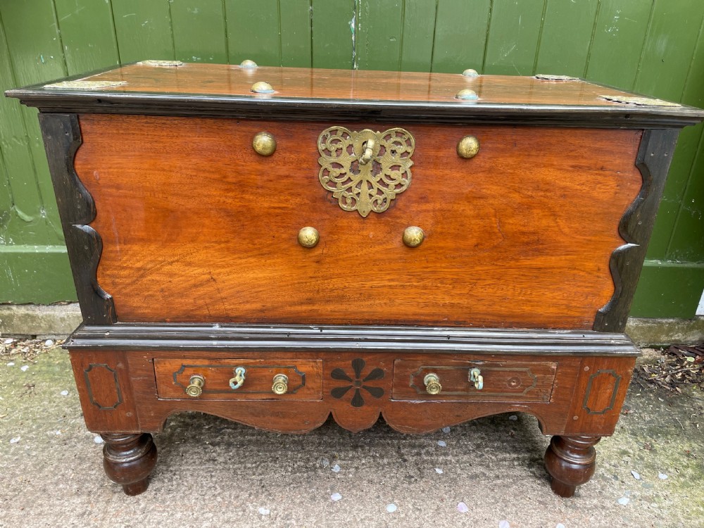 A 19th-century Dutch colonial teak and ebony chest with a pierced brass escutcheon, mounted on a two-drawer base with turned feet. View X