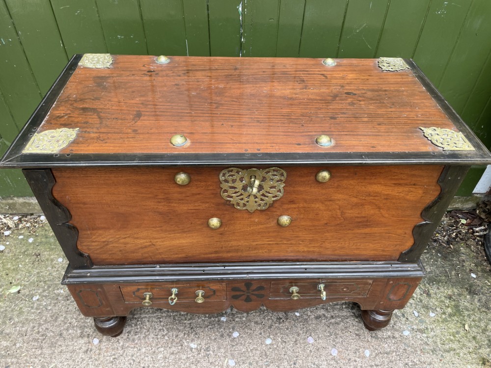 A 19th-century Dutch colonial teak and ebony chest with a pierced brass escutcheon, mounted on a two-drawer base with turned feet. View XIX